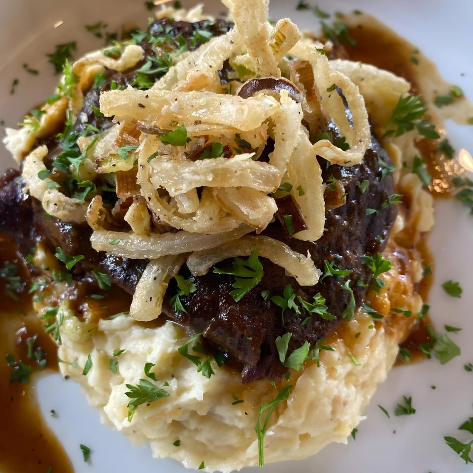 Braised short rib with gravy, mashed potatoes, crispy fried onions, and parsley on a white plate.